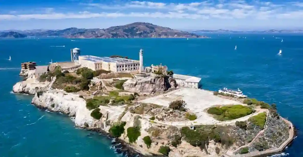 Donald Trump frente a un micrófono anunciando la reapertura de la prisión de Alcatraz, con vistas a la isla y la costa de San Francisco al fondo.